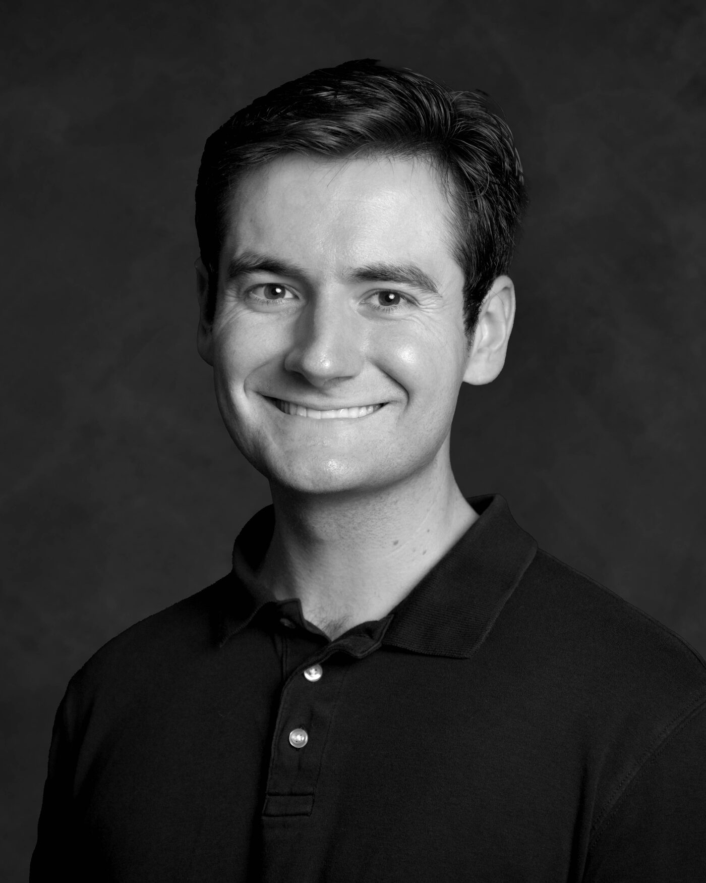 Black and white portrait of Owen Wolfe smiling, in a dark polo shirt against a dark backdrop
