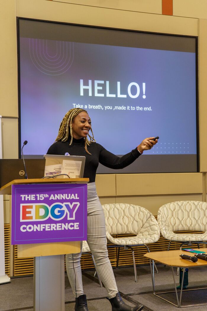 Speaker presenting beside an EDGY Conference podium sign, a HELLO slide on screen behind her