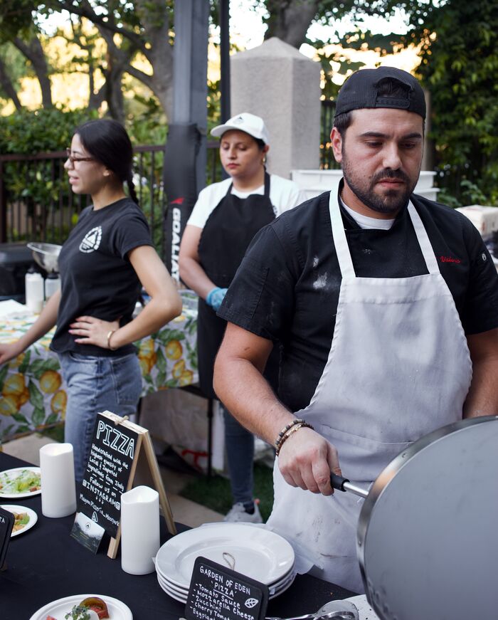 Chef in a white apron cooking at an outdoor catering station while staff prepare dishes