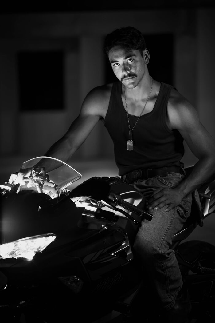 Black and white portrait of a man in a tank top and dog tags seated on a motorcycle, looking at the camera