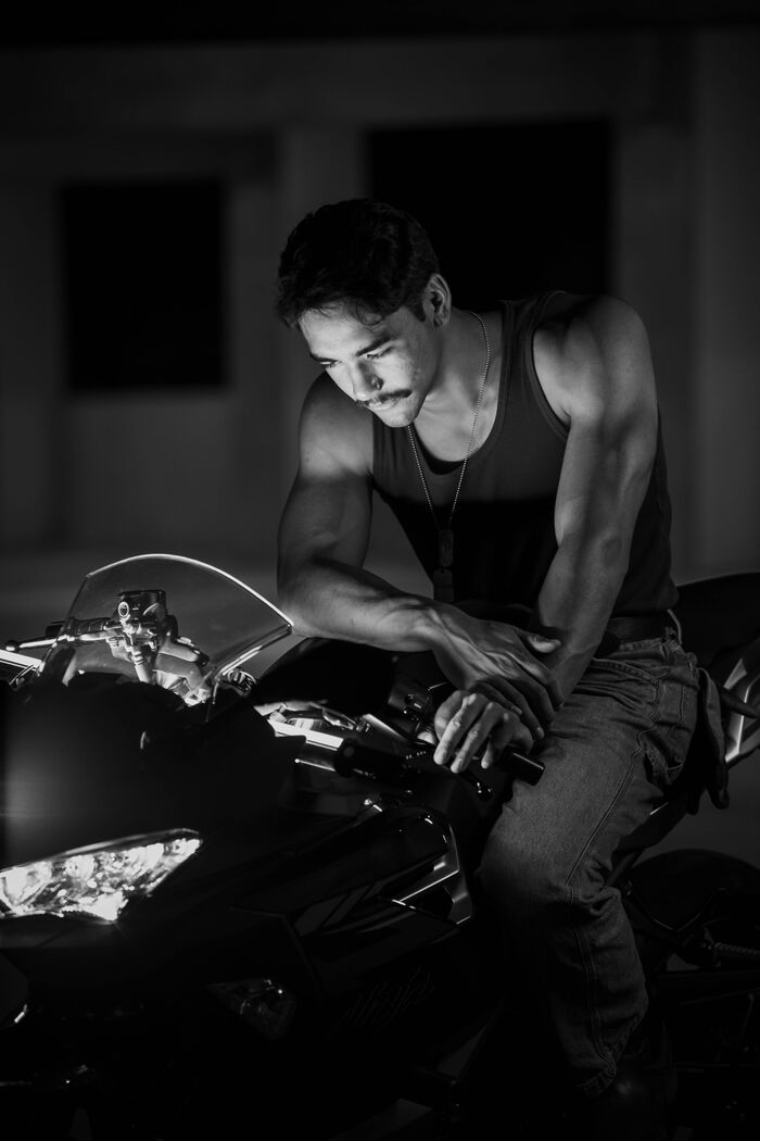 Black and white portrait of a man in a tank top leaning over a motorcycle, looking down