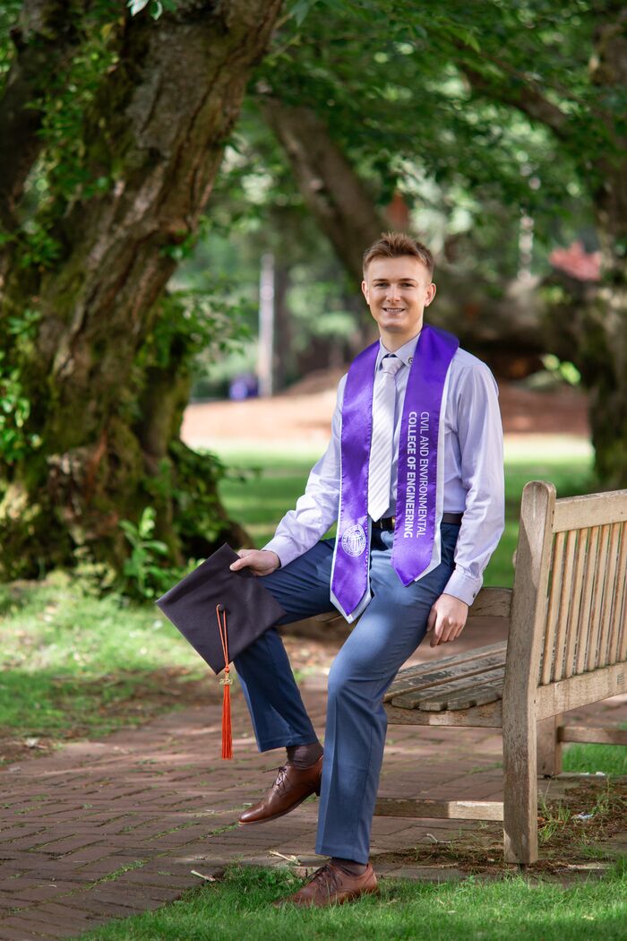 Smiling graduate in a purple stole sitting on a campus bench under trees, holding his cap