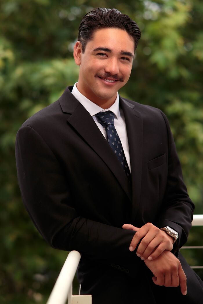 Man in a suit leaning on a white railing with trees behind him