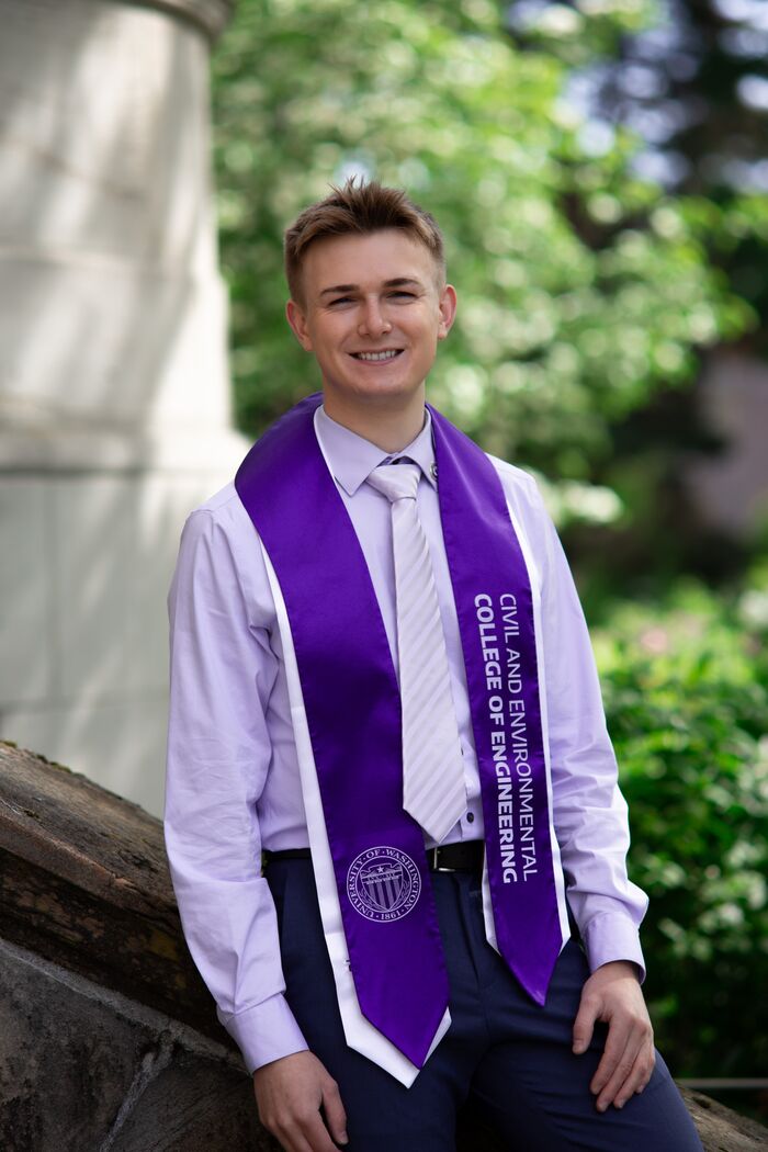 Smiling graduate in a lavender shirt and purple University of Washington stole, leaning on a stone wall