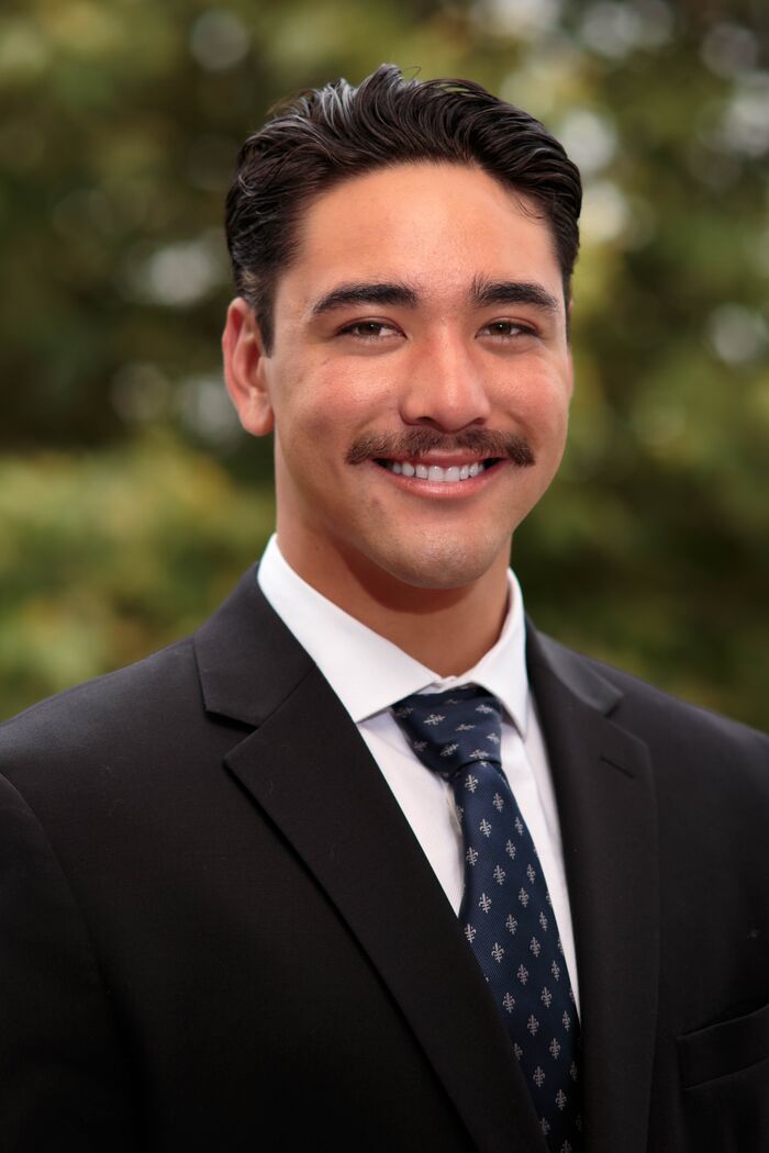 Smiling man in a dark suit and navy patterned tie against soft green foliage