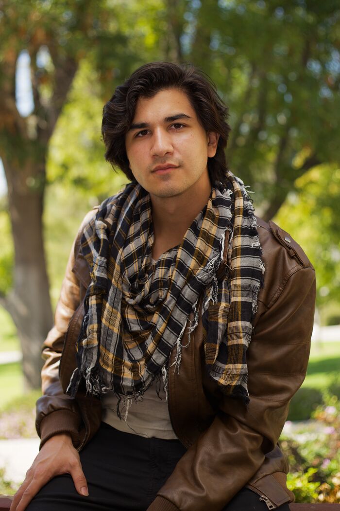 Young man in a leather jacket and plaid scarf seated outdoors in natural light