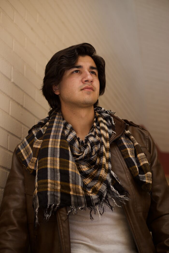 Young man in a leather jacket and plaid scarf leaning against a brick wall in warm light