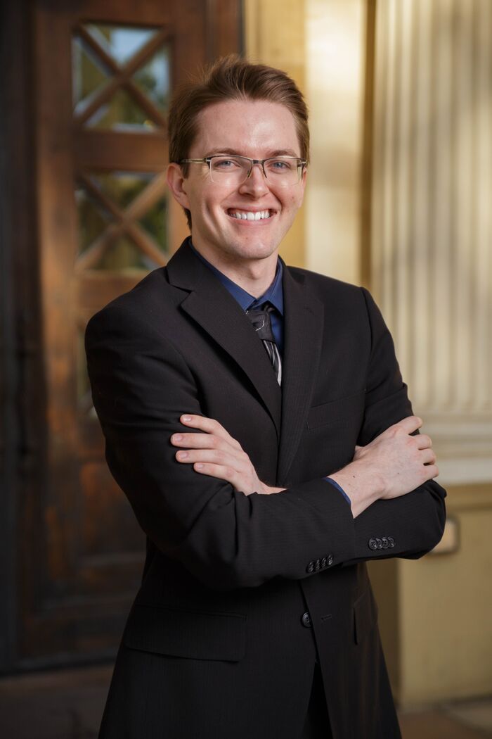 Smiling man in glasses and a dark suit, arms crossed, in a sunlit doorway