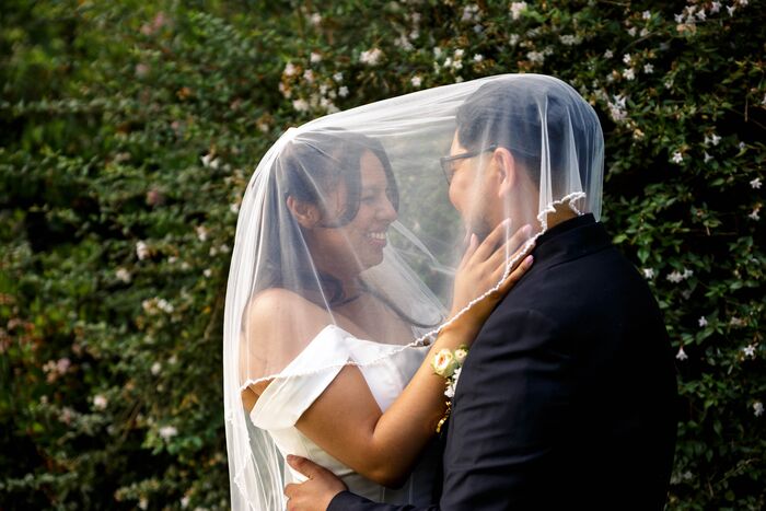 Bride and groom under her veil, smiling at each other in front of green foliage