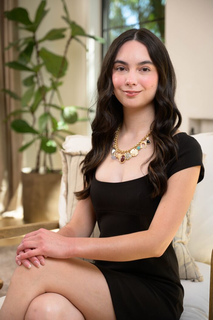 Young woman with dark hair in a black dress and gold necklace, seated