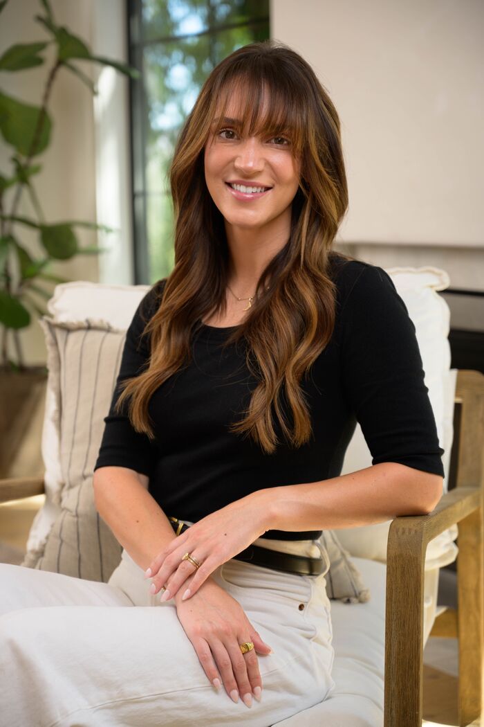 Woman with bangs in a black top and white trousers, seated