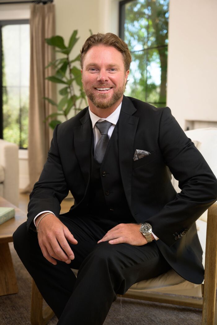Smiling man in a black three-piece suit seated in a sunlit room